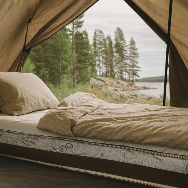 Camping mattress with beige bedding inside a canvas tent, partially uncovered to show the SlaapTyd logo, with a scenic view of pine trees and a lake visible through the open tent flap